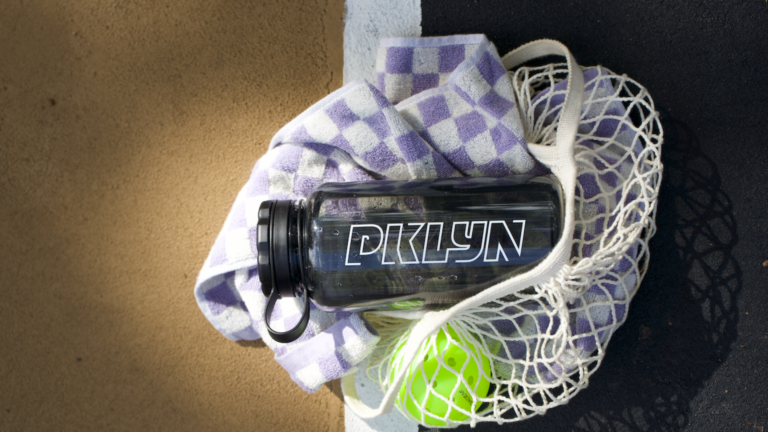 A PKLYN water bottle and green pickleball atop a purple and white checkered blanket